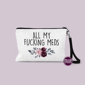 May include: A white canvas makeup bag with a black zipper and a black strap. The bag features a floral design with pink and burgundy flowers and the text "ALL MY FUCKING MEDS". The bag has a small logo in the bottom right corner that says "PLUM FOOLERY GIFT COMPANY".