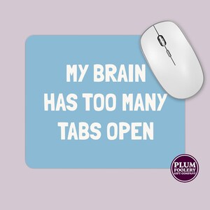 May include: A white wireless computer mouse sits on a light blue mouse mat. The mat has rounded corners and the text "MY BRAIN HAS TOO MANY TABS OPEN" in white. A Plum Foolery Gift Company logo is in the bottom right corner.