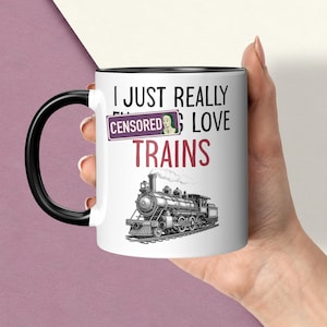 May include: A white ceramic mug with a black handle and rim. The mug features a black and red graphic of a steam locomotive train and the text I JUST REALLY LOVE TRAINS, with a purple rectangular CENSORED label covering part of the text.