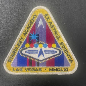 May include: A triangular embroidered patch featuring a yellow border with the text STARFLEET ACADEMY, EX ASTRIS, SCIENTA, LAS VEGAS, and MMCLXI. The center displays a white delta symbol, a blue star, and a saucer shape with colorful circular accents.