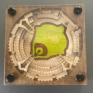 Op de afbeelding: Een houten onderzetter met een gedetailleerd lasergesneden ontwerp van een honkbalstadion. Het stadion is omgeven door een bruin houten frame met zwarte metalen accenten. Het veld is groen met een bruin binnenveld en een wit honkballogo in het midden.
