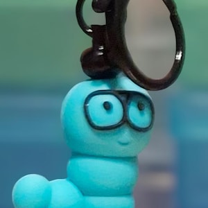 May include: A blue worm-shaped keychain with black glasses and a smiling face. The worm is perched on a small book with white pages. The keychain has a black clasp and a dark brown base.