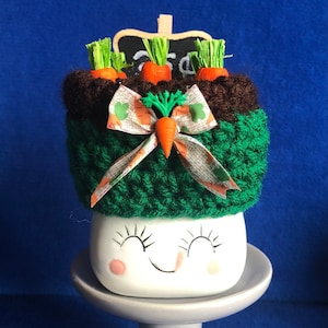 May include: A whimsical ceramic container with a smiling face, topped with a green and brown knitted cover. Miniature carrots and a bow adorn the top. The container sits on a gray pedestal, perfect for Easter decor.