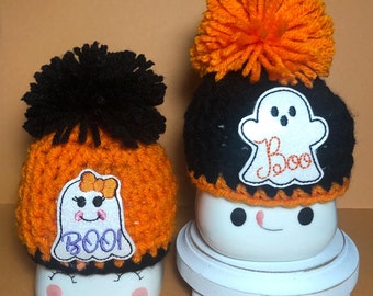 May include: Two crocheted Halloween hats with orange and black yarn. Each hat has a white ghost applique with the word "Boo!" embroidered on it. The hats are sitting on top of white ceramic mugs with painted eyes and a smile.