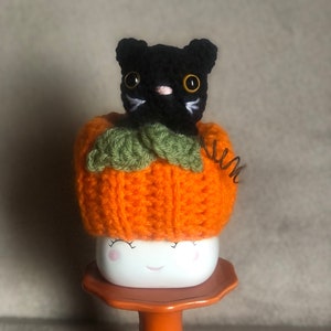 May include: A black crocheted cat wearing a crocheted orange pumpkin hat sits on a white mug with a smiling face. The mug is on an orange cake stand.
