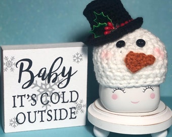 May include: A white crocheted snowman mug cozy with a black top hat, red berries, and green holly. The cozy is sitting on a white pedestal with the text "Baby It's Cold Outside" on a white wooden sign behind it.