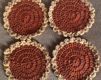 May include: Set of four crocheted coasters in a rust orange color with a cream colored border. The coasters are round and have a textured design.
