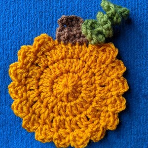 May include: A crocheted pumpkin coaster in orange with a brown stem and green leaves.