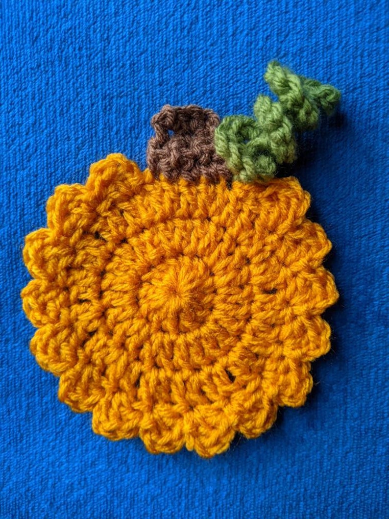 May include: A crocheted pumpkin coaster in orange with a brown stem and green leaves.