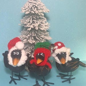 May include: Three black crow figurines wearing festive winter hats. One crow wears a red and white Santa hat, another wears a red and green crocheted hat, and the third wears a red and white Santa hat with snowflakes. The crows are standing in front of a small, snow-covered Christmas tree.