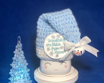 May include: A white ceramic marshmallow with a blue crocheted hat, a white and blue embroidered patch that says "Snowflakes are kisses from Heaven", and a small snowman charm. The marshmallow is sitting on a white wooden base.