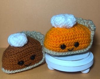 May include: Two crocheted pumpkin pie plush toys with smiling faces. The pie on the left is brown with a white whipped cream topping. The pie on the right is orange with a white whipped cream topping. The orange pie is sitting on a white pedestal.