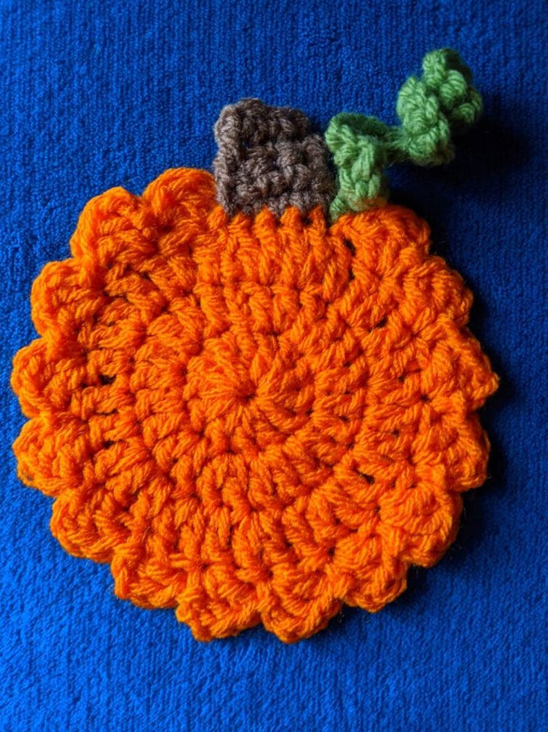May include: A crocheted orange pumpkin with a brown stem and green leaves. The pumpkin is on a blue background.