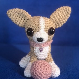 May include: A crocheted chihuahua toy with brown and white fur, sitting with a pink crocheted ball. The toy has black eyes and a pink collar with a purple gem.