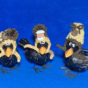 Puede incluir: Tres figuras de cuervos negros con picos naranjas, cada una con un gorro de ganchillo en tonos beige y marrón. Los gorros presentan pequeños detalles decorativos, incluyendo un icono de taza de café y un lazo marrón, con pompones texturizados.