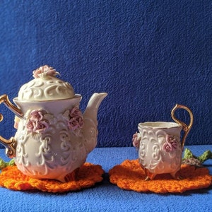 May include: A white porcelain teapot and teacup with pink floral accents and gold trim. The teapot and teacup are sitting on orange crocheted doilies.