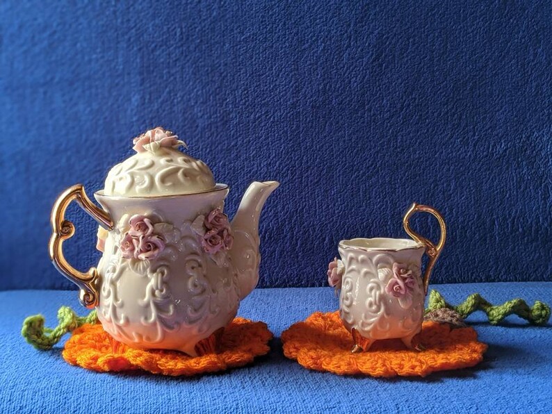 May include: A white porcelain teapot and teacup with pink floral accents and gold trim. The teapot and teacup are sitting on orange crocheted doilies.