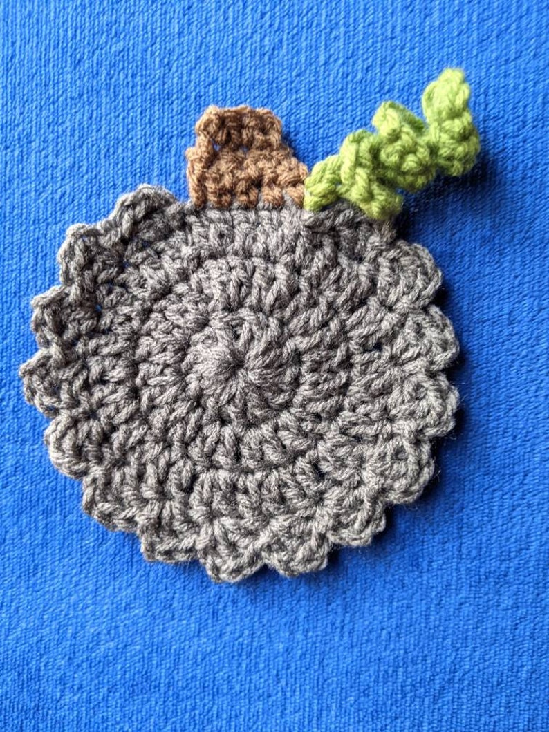 May include: A gray crocheted coaster with a brown crocheted stem and a green crocheted leaf on top.