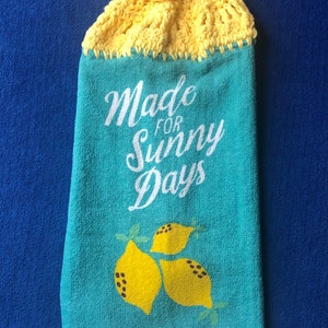 May include: A hanging kitchen towel with a yellow crocheted top and button closure. The turquoise towel features the text "Made for Sunny Days" and a lemon graphic. The bottom edge is white.