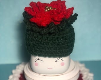May include: A white ceramic mug with a smiling face, wearing a green and red crocheted poinsettia hat with gold beads. The mug is sitting on a white crocheted doily with a red center.