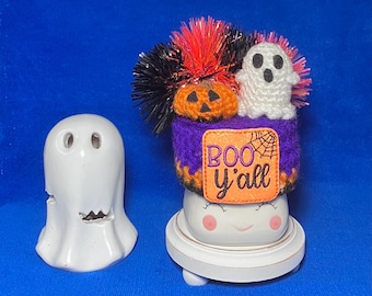 May include: A white ceramic ghost figure and a Halloween-themed decoration. The decoration features a purple crocheted base with an orange patch that says "BOO y'all". It is topped with a crocheted pumpkin, a white ghost, and orange and black tinsel.