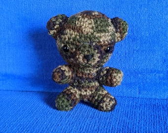 May include: A small crocheted teddy bear with a camouflage pattern in shades of green, brown, and beige. The bear has black button eyes and a black nose.