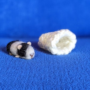 May include: A black and white needle felted guinea pig figurine laying on a blue surface next to a white crocheted sleeping bed.