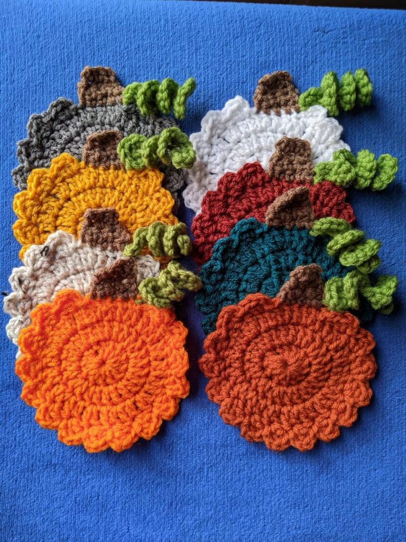 May include: Ten crocheted pumpkin coasters in various colors, including orange, red, green, white, and brown. Each coaster has a small green stem on top.