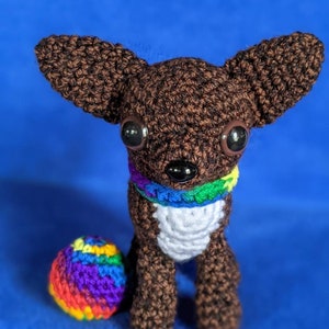 May include: A handmade crochet chihuahua dog with large, dark eyes and a rainbow-colored collar. The dog is brown with a white chest and is next to a small, rainbow-colored ball. The background is a solid blue.