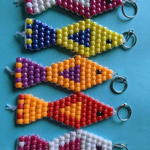 May include: Five colorful beaded fish keychains with silver key rings. The fish are made of small, round beads in various colors, including pink, white, yellow, blue, orange, and purple.