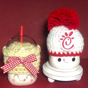 May include: Two white ceramic mugs with smiling faces, one wearing a red and white crocheted hat with a pom-pom and a red Chick-fil-A logo, the other wearing a yellow crocheted hat with a red and white gingham bow.