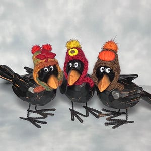 May include: Three black decorative crow figurines wearing colorful knit hats. The hats are adorned with pom-poms, a sunflower, and a pumpkin. The crows have wooden beaks and wire feet, perfect for fall or Halloween decor.