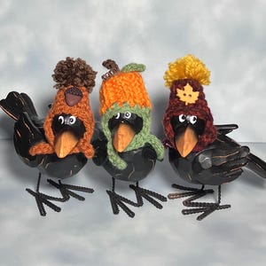 May include: Three black metal crow figurines, each wearing a different knitted hat. One has a brown acorn hat, another an orange pumpkin hat, and the third a burgundy hat with a yellow pom-pom. The crows have orange beaks and wire legs.