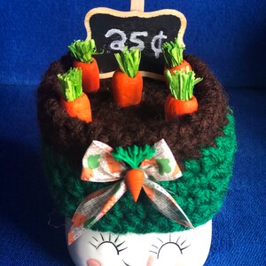 May include: A whimsical ceramic container with a smiling face, topped with a knitted brown and green section with miniature orange carrots. A small chalkboard sign displays "25¢". A decorative bow with a carrot accent adds a playful touch.