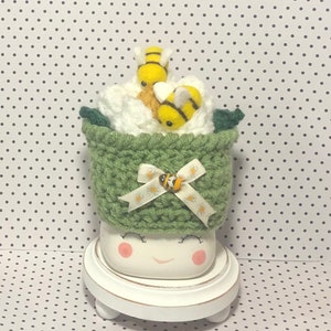 May include: A white ceramic mug with a green crocheted hat and a white ribbon with a bee design. The hat is filled with white felt and two yellow felt bees. The mug is sitting on a white pedestal.