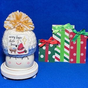 May include: A white ceramic mug with a smiling face, wearing a white and blue knit hat with a yellow and white pom-pom. A square patch on the hat shows a nativity scene with the text "every knee shall bow". Three wooden gift boxes sit nearby.