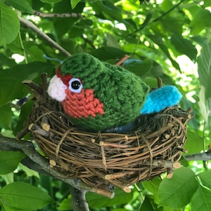 May include: A crocheted green and red bird with a blue tail sitting in a brown twig nest.