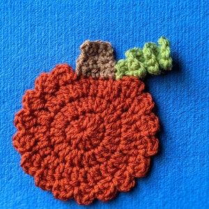 May include: A crocheted pumpkin applique in shades of orange, brown, and green. The pumpkin is round with a stem and a leaf.