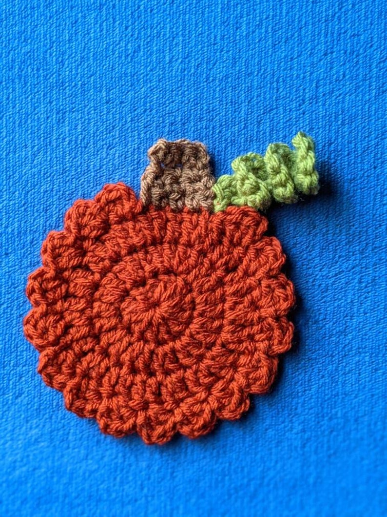 May include: A crocheted pumpkin applique in shades of orange, brown, and green. The pumpkin is round with a stem and a leaf.