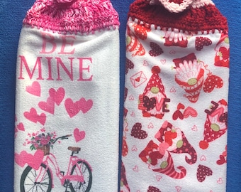 May include: Two Valentine's Day themed kitchen towels with crocheted toppers. The towel on the left has a pink topper and features a bicycle with a basket of flowers and the text "Be Mine". The towel on the right has a red topper and features a pattern of gnomes and hearts.