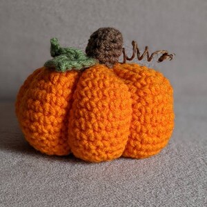 May include: A crocheted orange pumpkin with a green leaf and brown stem. The pumpkin is sitting on a gray surface.
