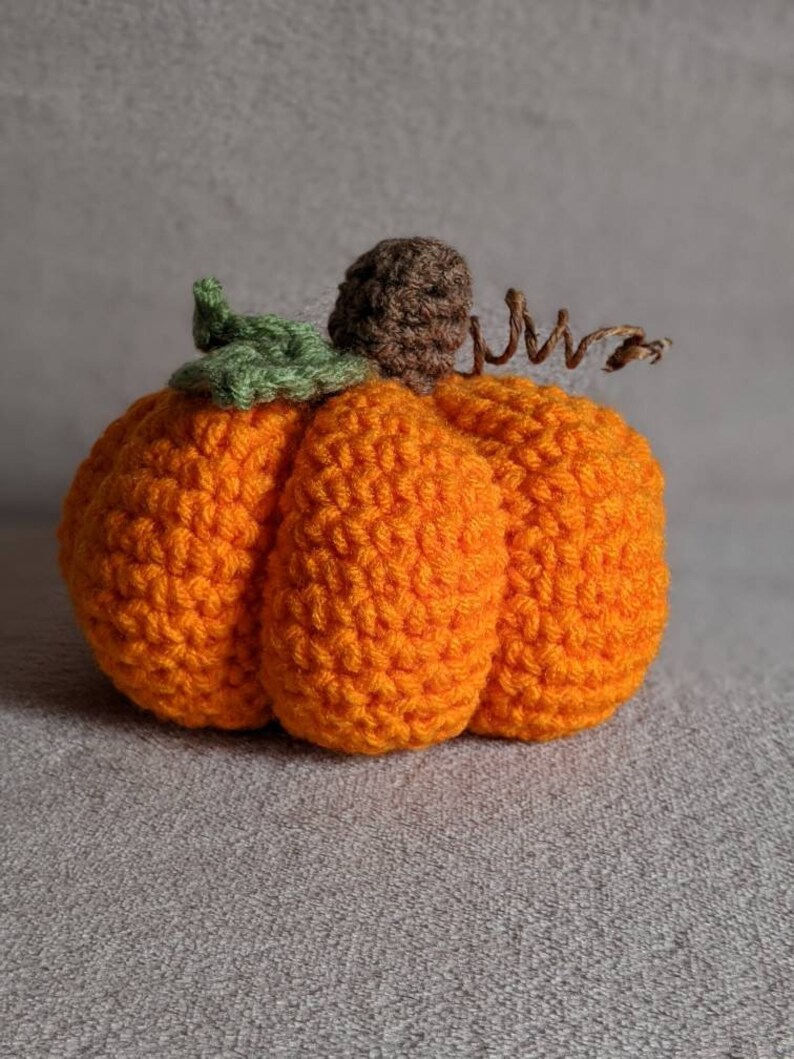 May include: A crocheted orange pumpkin with a green leaf and brown stem. The pumpkin is sitting on a gray surface.