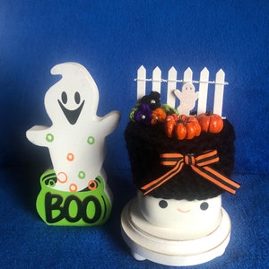 May include: A white ghost figurine with a green cauldron that says "BOO" and a black and white crocheted cauldron with a white picket fence, orange pumpkins, and a black bow.