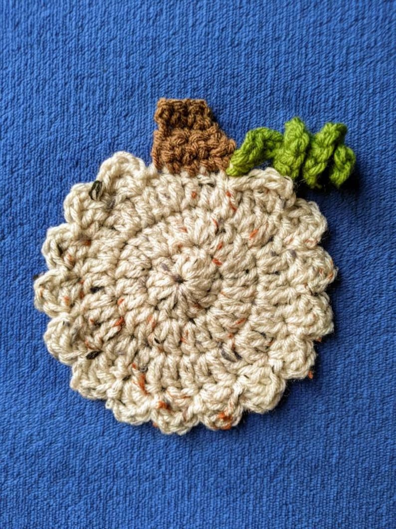 May include: A crocheted pumpkin with a brown stem and green leaves. The pumpkin is beige with brown and orange flecks.