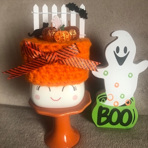 May include: Orange and black Halloween themed mug with a crocheted hat, a white picket fence, pumpkins, and a black cat. The mug is on a small orange pedestal. A white ghost with green and orange polka dots and the word "BOO" is on the right side of the image.