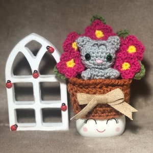 May include: A gray crocheted kitten sits in a brown crocheted pot with pink and yellow flowers. The pot is sitting on a white mug with a smiling face.