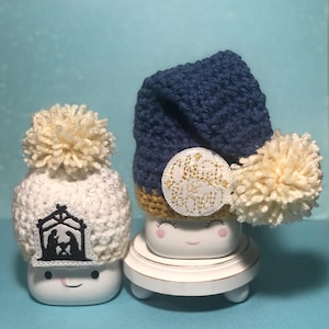May include: Two white ceramic mugs with smiling faces, wearing crocheted hats. One hat is white with a black nativity scene design and the other is blue with a gold "Christ is Born" patch.