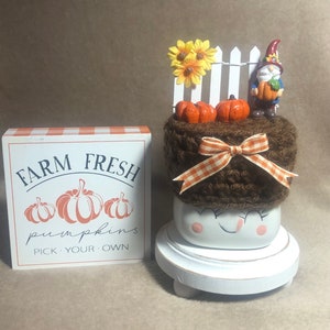 May include: A white ceramic mug with a brown knitted hat, a white picket fence, sunflowers, and a gnome holding a pumpkin. The mug has a smiling face and pink cheeks. The mug is sitting on a white wooden base. The sign reads "Farm Fresh Pumpkins Pick Your Own".