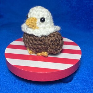 May include: A crocheted bald eagle figurine with a white head, brown body, and yellow beak and feet. The eagle sits on a round, red and white striped base, reminiscent of the American flag. The background is a solid blue.