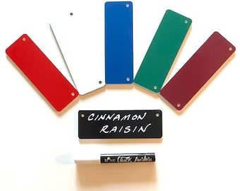 May include: A set of six rectangular tags in red, white, blue, green, and maroon, plus one black tag with the white text CINNAMON RAISIN. Each tag features small holes at the ends. A white chalk marker is positioned below the black tag.